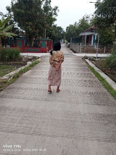 A person is seen from behind, walking away from the viewer along a narrow, paved concrete path in what appears to be a village or residential area. The individual is wearing a patterned long-sleeved top, a long skirt, and a black head covering (hijab), with sandals on their feet. Their hands are clasped behind their back as they walk.
The concrete path has distinct horizontal textured patterns across its surface. On either side, narrow drainage ditches are bordered by green grass and vegetation. To the left, a property enclosed by a red and green fence features several large trees, including a prominent banana plant. To the right, houses with various architectural styles, including tiled roofs, are visible behind fences, interspersed with more trees and greenery. The path recedes into the distance, flanked by similar structures and foliage.
The scene is lit by soft, diffused light, consistent with the early morning. A watermark in the bottom left corner of the image reads "vivo Y19s Akhtar & Arvan 14 Des 2025, 05.38," indicating the photo was taken at 5:38 AM on December 14, 2025. The overall setting, including the architecture and vegetation, strongly suggests a location in Indonesia, aligning with the context of Wanguk.