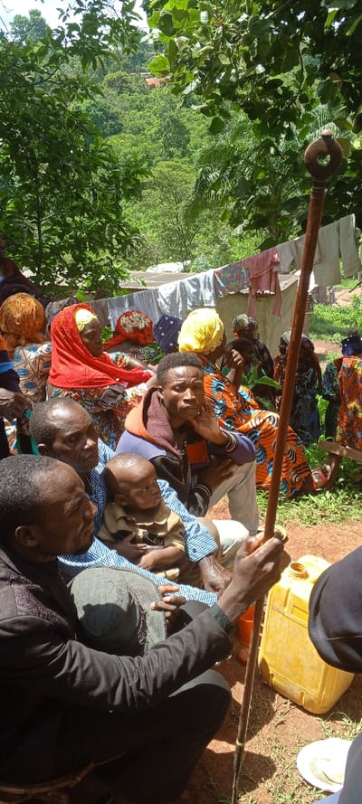 A group of people are seated outdoors, likely in a rural setting. There are many people sitting, some of them are wearing colorful dresses and head coverings. A small child is being held by a man in a blue checkered shirt. Another man is holding a tall walking stick. A yellow jerrycan sits nearby. Clothes are hanging on a line in the background. The surrounding environment appears to be lush and green.
