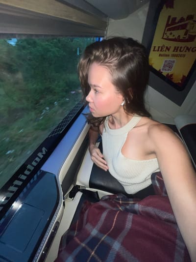 The image captures a young female individual seated inside a moving vehicle, likely a bus or train, with a window view of blurred green foliage. The person, positioned in the foreground, has long brown hair and is wearing a light-colored, possibly off-white, knitted tank top. A white earbud is visible in her left ear. Her face is shown in profile, gazing downwards and outwards through the window, suggesting observation of the passing scenery or contemplation.

The vehicle's interior features a comfortable-looking seat with dark upholstery and cream or white trim. A plaid blanket, featuring shades of red, blue, and yellow, is draped beside her, indicating a cozy travel environment. Below the window, there are dark blue or black panels with white stitching, and a partially visible, stylized word that appears to be "Widom" or similar, possibly a brand name, but it is reversed in the image.

Above the person's head, a yellow sign is prominently displayed with clear text and a QR code. The text reads "LIÊN HƯNG" and "Hotline: 19002579". This text, combined with the location context of Thôn 18, Vietnam, confirms the setting as a Vietnamese transport service. The lighting suggests natural daylight outside, possibly late afternoon or an overcast day, with softer ambient light inside the vehicle. The overall scene depicts a moment of travel and quiet observation.