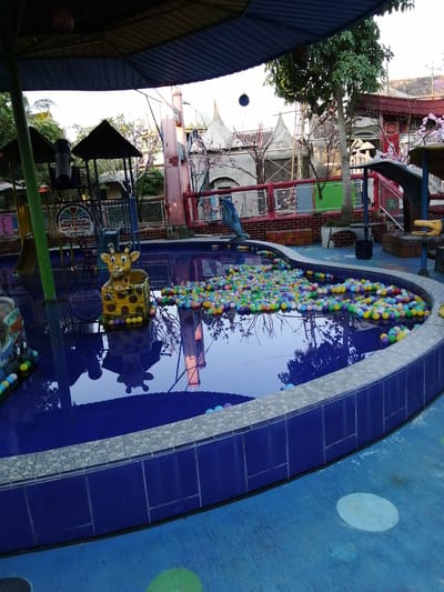 A high-angle, full shot captures a colorful ball pit situated in an outdoor swimming pool area. The pool, with its deep blue water, is partially filled with a multitude of plastic balls in various vibrant colors, including yellow, green, blue, pink, and orange. A cheerful, cartoonish yellow giraffe statue with large, expressive eyes and black spots sits prominently in the middle of the ball pit, appearing as if it's guarding the colorful collection. The pool's edge is lined with blue tiles and a lighter, pebble-like stone trim. Beyond the pool, a playground structure with a blue slide and red fencing is visible. Trees with green foliage and some pink blossoms are scattered throughout the scene. In the background, buildings with pitched roofs, some appearing to be themed or decorated with architectural elements, are present. The overall environment suggests a recreational or amusement park setting, likely designed for children. The location is identified as Kebonsari, Indonesia.