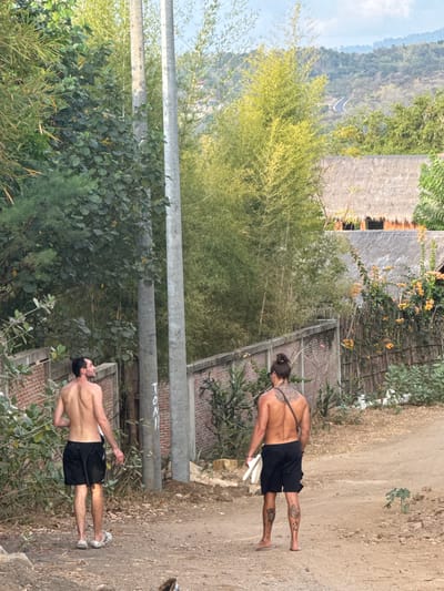 Two bare-chested men are walking on a dirt path in a rural setting. The man on the left wears black shorts and sandals, and the man on the right wears black shorts and carries a pair of white sandals. Both men have tattoos. The environment includes lush green vegetation, bamboo, a brick wall, two utility poles with writing on one, and thatched-roof buildings in the background. In the distance, a winding road is visible on a hillside. The scene depicts an outdoor environment in what appears to be a tropical or sub-tropical region, possibly within or near the area of Benete, Indonesia, suggested by the vegetation and building styles.