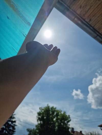 The image shows a low-angle view of a person's arm and hand reaching towards the sun in a partially cloudy blue sky. Part of a blue corrugated plastic roof is visible in the lower-left, with a wooden structure above it. Tree foliage is present in the upper-right. The location of Byala Slatina, Bulgaria is not determinable from the image.