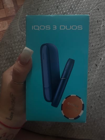 The image shows a hand holding an IQOS 3 DUOS product box. The box is blue and features the IQOS logo and "3 DUOS" text. Inside the box are two blue IQOS devices, a holder and a pocket charger. The hand holding the box has long, white-tipped fingernails and a silver ring on one finger. A tattoo reading "Love Yourself" is visible on the hand. The background is a textured gray surface, possibly a blanket or carpet. A circular orange sticker with Cyrillic text is on the lower right of the box. The scene is likely indoors, and the lighting suggests it could be daytime. There is no information in the image that places it specifically in the city of Razvedchik, Russia.