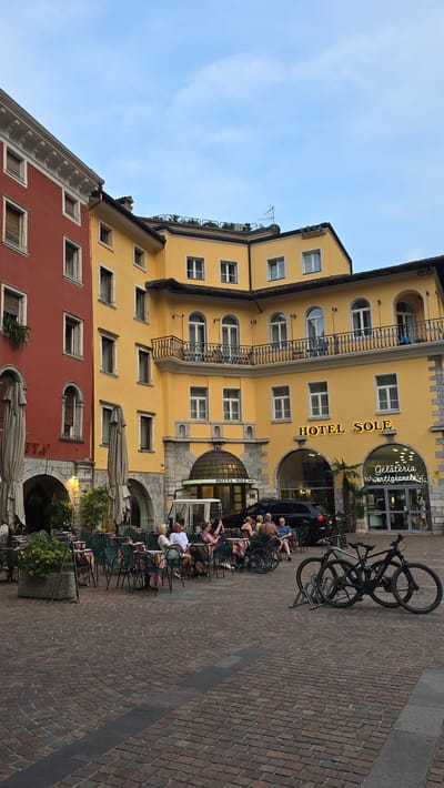 The image captures a serene evening scene in Riva del Garda, Italy, specifically in the Piazza Catena. The dominant structures are the red building on the left and the yellow Hotel Sole building on the right, both exhibiting classic Italian architecture with multiple stories, arched windows, and balconies. The sky is a soft blue with hints of evening light.

In the foreground, a cobblestone plaza is laid out, with several tables and chairs set up outside what appears to be a cafe or restaurant. People are seated at these tables, engaged in conversation and enjoying the ambiance. One individual is seated in a wheelchair. Two bicycles are parked in a bike rack to the right of the seating area, suggesting a mode of transportation for visitors.

The Hotel Sole building features a prominent sign and an entrance. Adjacent to it is a gelateria with glowing signage. The overall atmosphere is tranquil and inviting, indicative of a pleasant evening in a European town. The lighting suggests it is late afternoon or early evening.