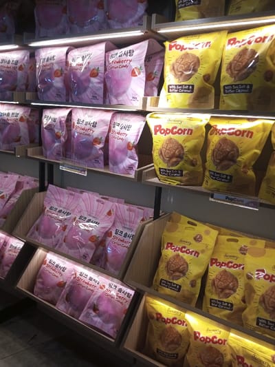 This image captures a static, eye-level view of retail display shelving inside a store located in Tân Kỳ Commune, Vietnam. The multi-tiered display unit is constructed from light-colored wood and dark metal supports, illuminated by linear LED lighting strips positioned beneath each shelf. The shelves are stocked with packaged snack foods. On the left side of the frame, several rows of pink stand-up pouches, labeled "chocomory Strawberry Cotton Candy" with accompanying Korean text ("핑크 솜사탕") and a stated weight of 70g, are neatly arranged both upright on shelves and within wooden display bins. Concurrently, on the right side, multiple rows of bright yellow stand-up pouches are displayed, identified as "chocomory Popcorn with Sea Salt & Butter Caramel." No individuals, active interactions, or movements are present in the scene.