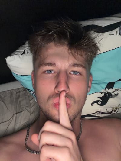 The image is a close-up selfie of a young man lying down, with his finger over his lips in a shushing gesture. He has light brown hair, blue eyes, and is wearing a silver chain necklace. He is lying on a pillow with a black and white cartoonish pattern. The background is dark. The image does not contain any information about the location in the city of Vanino, Russia.