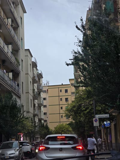 The image depicts a street scene in Salerno, Italy, on a cloudy day. In the foreground, the taillights of a silver Opel Corsa are illuminated, indicating it is stopped in traffic. The car has a license plate that appears to read "ED 829FX". To the left, another silver car, possibly a Nissan Micra with a similar license plate format, is also visible. Further ahead, a white SUV is partially obscured.

The street is lined with multi-story buildings. On the left, a pale yellow apartment building with numerous balconies is prominent. Across the street, another apartment building in a similar pale yellow hue is visible, featuring green trim. A large tree with dense green foliage partially obstructs the view of the buildings.

A man on a bicycle, wearing a white t-shirt and black pants, is in the middle of the road, facing away from the camera. Behind him, a few pedestrians are walking on the sidewalk. A street sign on a pole reads "Villamarina Isabella" and shows parking regulations. Neon signs are visible on the storefronts in the background. The overall atmosphere suggests a typical urban environment with moderate traffic.