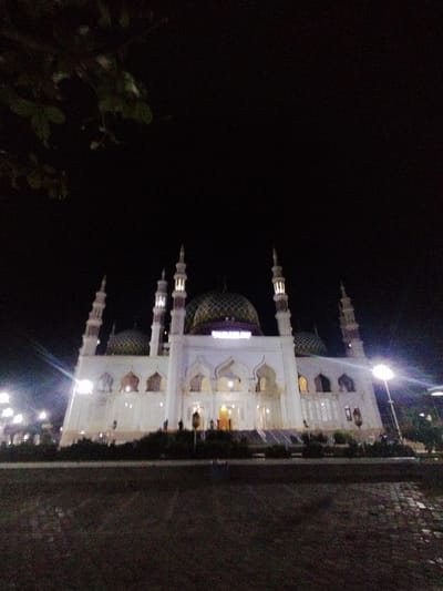 The image displays a brightly illuminated mosque at night in Kotapantonlabu, Indonesia. The central subject is a large, white mosque featuring multiple minarets with lighted upper sections and a prominent central dome along with several smaller domes, all appearing to have a patterned surface of green and gold. A sign above the main entrance of the mosque is discernible, likely reading "MASJID RAYA". The mosque's architecture includes multiple arched windows and doorways on its facade. Artificial lights surrounding the structure create significant lens flares, particularly on the left and right sides of the frame, contrasting sharply with the dark, clear night sky above. The foreground consists of dark, textured ground, possibly a paved plaza or roadway, separated from the mosque by a line of dark, indistinct foliage. A tall streetlamp with a bright light is visible on the right side of the frame, contributing to the overall illumination. Several indistinct human figures are sparsely visible near the base of the mosque and on the adjacent grounds.