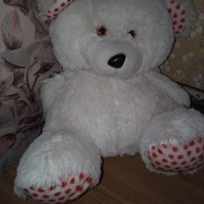 A white stuffed teddy bear with red hearts on its ears and paws sits on a wooden surface. The bear's eyes are brown, and its nose is black. The background is a patterned wallpaper. The image was taken in Slobidka, Ukraine.