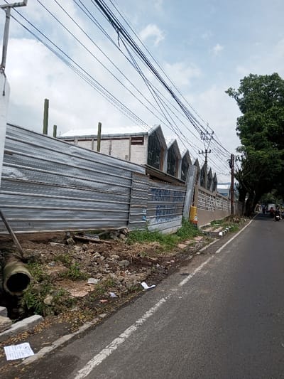 The scene depicts an outdoor urban environment in Khajuripada, India, on a clear day with scattered clouds. A two-lane asphalt road runs from the mid-right foreground into the background, with a solid white line marking its right edge. To the left of the road, a narrow dirt shoulder separates it from a temporary construction barrier made of corrugated metal panels. This barrier is partially covered in graffiti, including blue text reading "TERIMA DONAS... GERBANG..." on its right section. Behind the barrier, a building with a light-colored facade is visible, topped by a series of three prominent, dark-roofed structures featuring sharply gabled roofs with vertical elements. Several wooden posts extend vertically from behind the barrier, suggesting ongoing construction or renovation.
Further to the left foreground, a concrete drainage pipe opens into a channel obscured by dirt and sparse vegetation. The ground between the road and the barrier is uneven, covered in dirt, rocks, grass, and scattered litter including discarded papers. Above the buildings, numerous dark utility cables crisscross the sky, supported by a wooden utility pole with multiple cross-arms positioned along the roadside. To the right of the road, a low concrete curb separates it from a light-colored concrete block wall with a decorative open pattern, behind which mature trees with dense green foliage line the street. In the distant background on the road, two individuals on motorcycles are observed traveling away from the viewer.