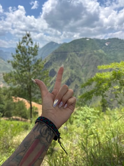 In an outdoor shot taken at Lũng Cú Commune, Vietnam, a person's left arm and hand are prominently featured against a backdrop of rolling green mountains and a partly cloudy sky. The hand is making a gesture, with the index and little fingers extended. The arm, which is tattooed, is adorned with a colorful braided bracelet. The mountainous landscape, partly obscured by trees, stretches into the distance under the cloudy sky.