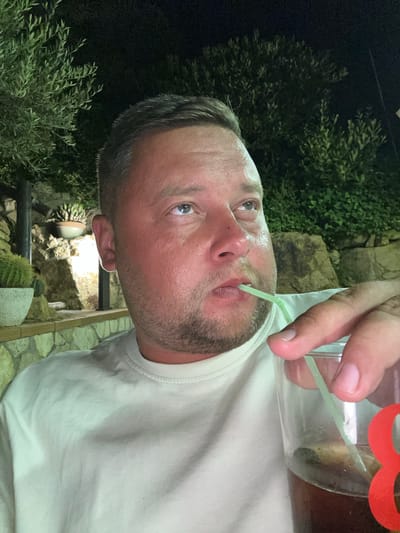The image shows a close-up, selfie-style shot of a man drinking from a glass with a green straw. He has fair skin, short brown hair, and is wearing a light-colored t-shirt. He appears to be outdoors at night, with dark foliage and what looks like stone wall construction visible in the background. The man is looking upwards and to his right, with his mouth slightly open as he sips through the straw. The glass contains a dark liquid with some ice. The scene is set in a dimly lit outdoor environment, possibly a restaurant or bar terrace in Sant Feliu de Guíxols, Spain, given the presence of Mediterranean vegetation.