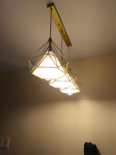 A linear chandelier with a gold-colored metal frame hangs from a light-colored ceiling. The chandelier has three pendant lights, each enclosed in a geometric, diamond-shaped cage. Inside the cages are white, translucent shades that diffuse the light from the bulbs. The light emitted is warm and bright, illuminating the surrounding area. The ceiling appears to be textured.

Below the chandelier, the wall is painted a warm beige color. On the left side of the wall, near the bottom, are two light switches. On the right side of the wall, at the very bottom, is a dark-colored object that appears to be a piece of luggage or a bin with a handle.

The scene is indoors, likely a room in a home or establishment. The lighting suggests it could be evening or a dimly lit space where the chandelier is the primary light source. There is no indication of the time of day or weather. No people are visible in the image. The overall impression is one of modern interior lighting. There is no visible text in the image.