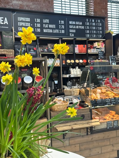 The image captures an interior view of a cafe or bakery located in Liepaja, Latvia, evident from the Latvian text on the menu board and other signage. The foreground is dominated by a large potted plant, primarily featuring vibrant yellow daffodils with long green stems and leaves, interspersed with some reddish-purple flowering plants. Several wooden sticks or stirrers protrude from the white pot containing the plants.
In the midground, a black menu board with white lettering is prominently displayed, listing various beverages and their prices. English entries include "ESPRESSO" (1.50, 2.60), "LATTE" (2.20), "FLAT WHITE" (2.70), and "MATCHA LATTE" (3.00). Latvian entries include "MELNA KAFIJA" (Black Coffee) at 1.50/1.60/2.20 (S/M/XL), "KAFIJA AR PIENU" (Coffee with Milk) at 1.70/2.00/2.60, "KAKAO" (Cocoa) at 1.80/2.20/2.70, and "TEJA" (Tea) at 1.00/1.30/1.50.
Below the menu board, a multi-tiered shelving unit, dark in color, is stocked with various retail products, including packaged coffee bags (e.g., "Colombia Brazile," "Gurmans Amaretto") and boxes labeled "KALVE." To the right, a white espresso machine and related equipment are visible on a counter, along with disposable cups. A person, dressed in a dark green or black top, is partially visible behind the counter, operating or standing near the coffee machine.
Further to the right, a clear glass display case presents an assortment of baked goods, including croissants, various pastries, and what appear to be donuts or buns. Small price tags in Latvian are visible within the display, with one partially legible as "Liepājas" (likely indicating Liepaja-specific item). Several woven baskets are present; one on a shelf contains small packaged items, and another on the counter holds more baked goods. A white digital scale and a red payment terminal are also positioned on the counter.
The background wall above the shelving features exposed reddish-brown brickwork. Below the counter, the structure is clad in light-colored wooden paneling. Horizontal blinds are visible through windows in the upper background, suggesting natural light sources.