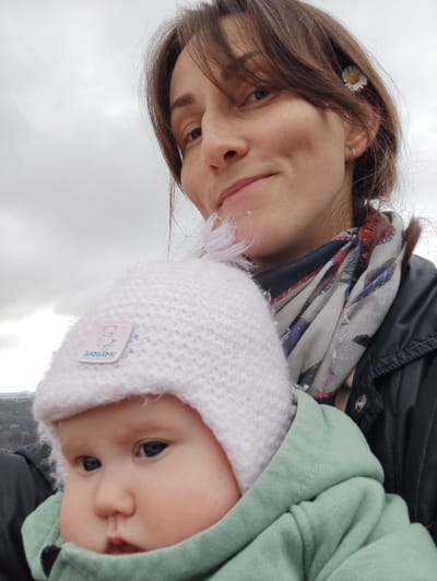 A close-up, chest-up selfie depicts an adult female holding an infant outdoors in Bestuzhevskoye, Russia, under an overcast sky with a blurred distant city visible in the background.

The adult female, positioned in the upper portion of the frame, has brown hair styled with bangs, light freckles on her nose and cheeks, and a faint smile. A small white daisy with a yellow center is tucked into her hair above her right ear, which also bears a small silver hoop earring. She wears a dark jacket, possibly black, and a multi-colored scarf featuring grey, black, red, and blue patterns around her neck.

The infant, held by the female in a front-carry position, occupies the lower portion of the frame. The infant has light skin, blue eyes, and a neutral expression, looking slightly to the left. The infant wears a light pink knitted hat with a white rectangular label featuring a blue teddy bear illustration and the text "BARRABAS" visible. A light green hooded garment, likely a jacket or hoodie, covers the infant's body.

The background consists primarily of a uniformly light grey, overcast sky. In the lower-left corner, a blurred landscape shows distant urban structures and patches of vegetation on elevated terrain, consistent with a city overview.