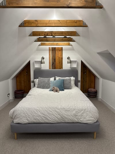 This image captures a tastefully decorated bedroom, likely an attic conversion or a room beneath a pitched roof, given the prominent sloped white ceilings. The room is dominated by a large bed featuring a grey upholstered headboard and frame. The bed is neatly made with a white quilted duvet and an arrangement of pillows in white, grey, and light blue, with a small, plush, light-colored toy resting on top.

A striking architectural feature on the left side of the room consists of several rustic wooden beams, each secured with visible metal connectors, extending vertically from the floor to the sloped ceiling. These beams add a raw, natural element to the otherwise clean, white walls. Behind the bed, a unique headboard accent is created by vertical wooden paneling in various warm tones.

Flanking the bed are two modern silver sconce lights mounted on the white walls, each positioned below a small, white square floating shelf. In the recessed areas on either side of the bed, formed by the ceiling slope, sit two round, woven baskets with striped patterns, adding texture and storage. The entire floor is covered in a soft, light grey carpet, enhancing the room's cozy and tranquil ambiance. The setting appears neat and well-maintained, suggesting a peaceful retreat. There is no visible text or human activity in the image.