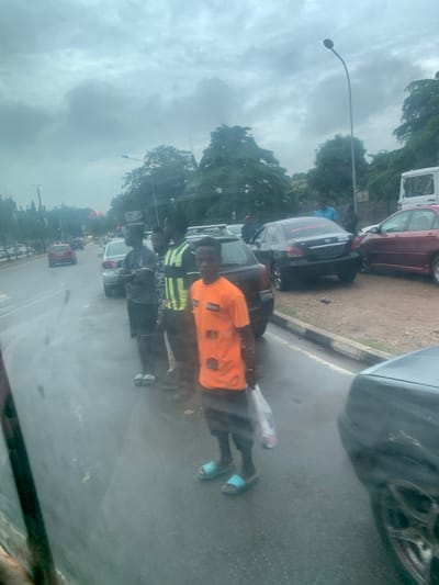 The image depicts a street scene in Aso, Nigeria, likely during a cloudy or overcast day, as indicated by the muted lighting and gray skies. A group of young men are standing by the side of a wet road, suggesting recent rain or the presence of puddles. They appear to be waiting, possibly for transportation or for a business opportunity.

In the foreground, a young man in an orange t-shirt and light blue flip-flops holds a white plastic bag. To his left, another young man wears a dark gray t-shirt and shorts, with white-striped flip-flops. Further down the line, a man in a high-visibility vest with green and yellow stripes is visible. The presence of multiple cars, including a black sedan with red lettering on the rear window, a red sedan, and other vehicles partially obscured, suggests moderate traffic. The road itself has white and yellow lane markings, and a concrete curb separates the road from the sidewalk area where the individuals are standing. Trees line the background, adding to the urban greenery. Streetlights are also visible, suggesting this is a developed area within the city. The overall atmosphere is one of everyday urban life, with a hint of the lingering effects of wet weather.