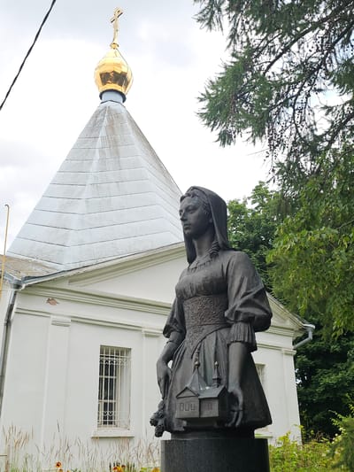 The image shows a bronze statue of a woman holding a miniature building in front of a white church with a golden dome. The statue is of a woman dressed in historical clothing, with her head covered by a veil and her hair styled in curls. She is wearing a long, flowing dress with intricate detailing on the bodice and sleeves. In her hands, she holds a small model of a church, complete with a dome and windows. The church in the background is a white, rectangular building with a stepped roof and a golden dome topped with a cross. A small, barred window is visible on the side of the church. The scene is set outdoors, with lush green trees and foliage surrounding the church and statue. The sky is overcast with a few wisps of clouds. This statue and church are located in the village of Ulitkino, Russia, a suburb of Moscow.