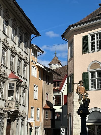 The image depicts a street scene in Schaffhausen, Switzerland, with buildings on either side angled towards the center. The sky is visible in the space between the buildings, featuring a clear blue hue with sparse clouds.

The buildings are of varying architectural styles, with one featuring ornate, classical detailing around the windows. The windows are primarily multi-pane, and some have green shutters. A fountain with a statue of a crossbowman is visible on the right, partially obscuring the view of a building behind it. The tower of a church or similar structure is visible further down the street between the buildings. A sign is visible to the right of the crossbowman.
