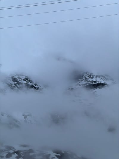 The image displays a high-altitude, mountainous landscape shrouded in dense mist or clouds, creating an atmospheric and somewhat obscured view. The photograph is oriented vertically, with the natural scene appearing rotated.

The primary subjects are rugged, dark mountain peaks partially covered with patches of white snow or ice. These peaks are largely enveloped by thick, diffuse grey and white clouds or fog, which reduces visibility and gives the scene a mysterious, vast quality. Portions of the mountains are visible as stark, dark masses contrasting with the lighter fog and snow.

In the upper left section of the image, several thin, dark lines, likely wires (possibly from a cable car, ski lift, or power lines), traverse diagonally across a lighter grey-blue sky, indicating an overcast day. These wires provide a sense of scale and human presence within the grand natural environment.

The weather is clearly overcast and foggy, limiting the panoramic view typical of mountainscapes but emphasizing the textures and forms that do emerge from the mist. There are no visible people or discernible activities taking place, and no visible text. The location context of Poschiavo, Switzerland, reinforces the depiction of a Swiss alpine setting, characterized by its dramatic mountain ranges and often challenging weather conditions. The overall mood is serene yet powerful, highlighting nature's grandeur even when partially concealed.
