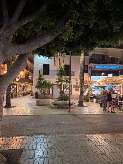 This image depicts a nighttime scene on a street in Sant Antoni De Portmany, Spain, likely a pedestrian walkway adjacent to a commercial area. In the foreground, a large tree trunk and branches dominate the left side, casting shadows across the patterned pavement. The pavement itself is composed of wavy, interlocking tiles, with a darker section of larger rectangular stones to the right.

Further into the scene, a white building with a dark-paned balcony stands prominently. Flags, one appearing to be the Catalan flag, are attached to the building. In front of this building, two large concrete planters hold spiky plants, likely yucca.

To the right of the white building, a restaurant or bar area is visible, illuminated by string lights and covered by a canopy. Tables and chairs are set up outside, with several people seated or standing. Two women are the most clearly visible individuals in this area, standing near a menu board. Another person, possibly an employee, is seen behind a counter or serving area.

Further down the street, more buildings with balconies and illuminated windows suggest shops and residences. A neon blue sign above one establishment reads "SNACK BAR LINARES". The overall atmosphere is that of a lively evening promenade in a Mediterranean town.