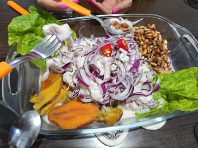 A close-up, high-angle shot shows a glass dish filled with ceviche, a Peruvian seafood dish, situated on a dark wooden surface. The ceviche consists of white fish marinated in citrus juices, adorned with thinly sliced red onions and garnished with two slices of red chili peppers. The dish also contains cooked sweet potato slices and toasted corn kernels. Two people are visible, one holding a fork with a piece of fish, and another holding a spoon, both likely involved in eating the dish. The environment suggests an informal dining setting in La Plata, Argentina.