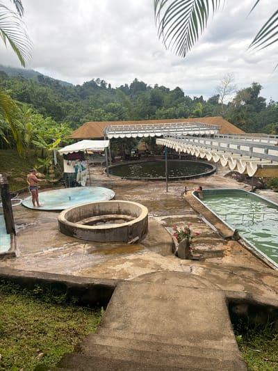 In Khanh Vinh, Vietnam, an outdoor recreational area features several pools. A man in swimming trunks stands by a shallow, circular pool with a fountain in its center, holding a hose. Adjacent to this is a larger, oval-shaped swimming pool filled with green water. Further in the background, a large, dark circular pool is visible beneath a covered walkway. The landscape is lush, with dense green foliage and trees covering the hillsides surrounding the area. The sky is overcast.