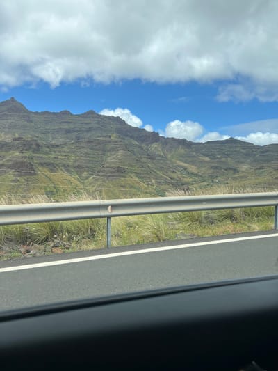 A view from a vehicle traveling on a roadway in Mogán, Spain. The window frame of the vehicle is visible on the right side of the image. A guardrail runs parallel to the roadway, separating the road from a steep mountain slope covered in green vegetation. The sky is blue with patches of white clouds. Power lines are visible against the mountain slope. The road surface is gray with a white dividing line.