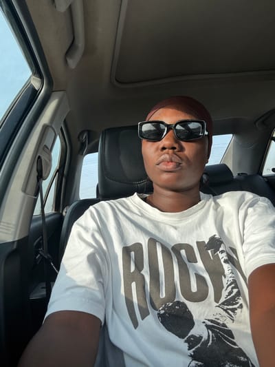 A dark-skinned person wearing a brown beanie and black sunglasses is sitting in a car. The person is wearing a white t-shirt with black writing and a black and white graphic. The car's interior is gray, and a seatbelt is visible. The image suggests the car is parked or stationary. There's no information about Kuchingoro, Nigeria.