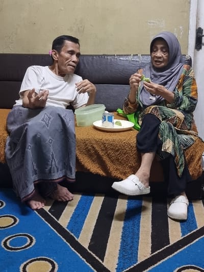 The image displays two elderly individuals, an Indonesian male and female, seated on a dark sofa with an orange-brown throw blanket, in an indoor residential setting in Pacet, Indonesia. The male, positioned on the left, wears a white short-sleeved t-shirt and a grey patterned sarong, with bare feet, and is captured mid-gesture with his right hand while looking towards the female. The female, situated on the right, wears a light grey hijab, a long-sleeved dress with a multi-colored abstract pattern, black trousers, and white closed-toe slip-on shoes with buckles, and is engaged in manipulating a small green object with both hands. Between the individuals on the sofa, a white plate holds a small metal can and a small green item, next to a light green plastic container with a translucent lid. The background consists of a light beige wall, and the foreground floor is covered by a patterned rug featuring blue, black, and beige stripes along with circular blue and yellow motifs.