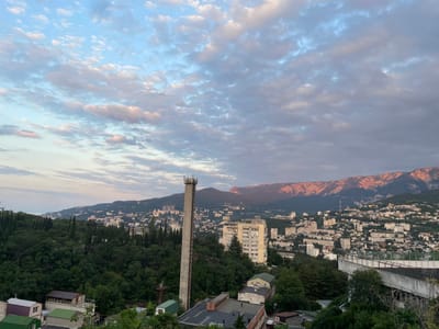 This is a panoramic, high-angle outdoor shot of the city of Yalta, Ukraine, under a partially cloudy sky. The foreground is dominated by lush green trees and the rooftops of several buildings. A tall, slender concrete tower with antenna equipment on top stands prominently in the mid-ground. Beyond this, the city sprawls across hillsides, characterized by a dense collection of buildings of varying heights and architectural styles, with some taller, modern structures interspersed among older, lower-rise buildings. In the far background, a mountain range is visible, its peaks catching the warm, rosy glow of sunset. The sea is faintly visible on the left horizon. The overall scene depicts an urban landscape nestled against a natural backdrop, under a twilight sky.