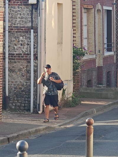 In Étretat, France, a person wearing a camouflage t-shirt, black shorts, a black baseball cap, and black sandals walks down a cobblestone street, carrying a black bag over their right shoulder. The person is holding a clear drinking glass in their right hand and appears to be walking towards the viewer. The background consists of stone and brick buildings, some with exposed brick and stone facades. A patch of green foliage with purple flowers is visible near one of the buildings. The street is lined with a concrete curb and a sidewalk. The sunlight casts long shadows across the street. A silver bollard is positioned in the foreground.