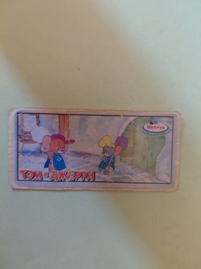 A rectangular sticker depicts cartoon characters Tom and Jerry. The characters are shown in a snowy outdoor scene. On the left is Jerry, wearing a red hat and blue garment. On the right is Tom, wearing a yellow hat and blue garment. In the upper right corner of the sticker is a logo with text that reads "Нептун" and "Мир развлечений и уюта". The sticker is affixed to a light-colored surface.