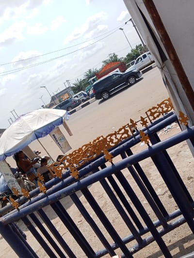 The image depicts a street scene in the city of Naze, Nigeria, captured from a slightly elevated and angled perspective. In the immediate foreground, a dark blue metal fence with ornate, gold-painted decorative elements spans the lower portion of the frame. Beyond the fence, a light brown dirt or sandy ground transitions to a paved asphalt road.
In the midground, to the left, several individuals are gathered under a white and blue umbrella. One person, wearing a brown garment, is visible near a motorcycle. A sign displaying "AVAILABLE" is partially legible, hanging from the umbrella's frame. On the road, traffic is moving; a dark-colored SUV (likely black or dark grey) proceeds leftward, followed by a white pickup truck. Further down the road, a large truck carrying a reddish-orange load is visible, along with additional vehicles including another dark SUV and a white van. Utility poles with streetlights and overhead power lines line the thoroughfare. The background features various buildings, suggesting commercial or residential structures, interspersed with green vegetation, including palm trees. The sky is bright with scattered white clouds, indicating daylight conditions.