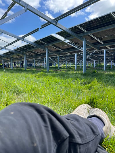 The scene depicts a ground-level view under a grid of solar panels, likely a solar farm. The location is suggested to be Grange Court, Ireland, though no specific landmarks confirm this. The panels are supported by a metal framework. Bright green grass grows under and around the structure. Visible in the foreground are the legs of a person wearing black trousers, gray socks, and worn light-colored sneakers, sitting in the grass. In the distance, near the middle of the frame, are figures who appear to be workers, one possibly on a ladder. The sky, with scattered clouds, is visible through gaps in the solar panel grid.