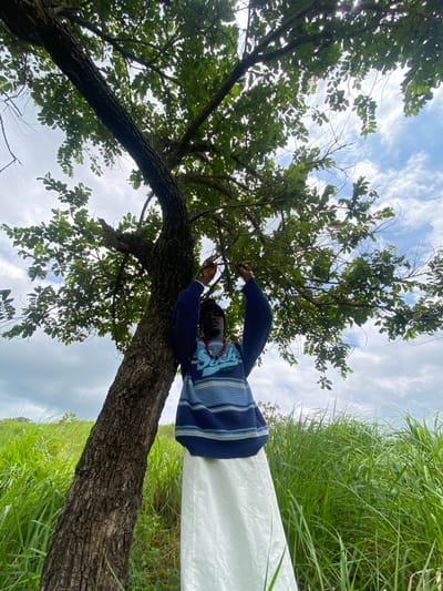A person stands leaning against the trunk of a large tree in a grassy field under a cloudy sky in Achara, Nigeria. The person is wearing a blue and white striped sweater and a long white skirt, with a red beaded necklace. Their left arm is raised, touching the branches of the tree. The field is covered in tall, green grass, and the tree's leaves are lush and green, obscuring parts of the sky. The overall scene suggests an outdoor, natural setting.