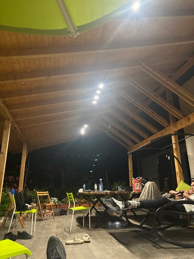 The image depicts an outdoor patio area at night, under a wooden roof structure. The scene appears to be a backyard or a covered porch. In the foreground, there are several chairs, including two bright green folding chairs and a wooden folding chair. One of the green chairs has a black backpack on it. A pair of black sneakers are on the ground near the green chairs, and a pair of light-colored slides are also on the floor.

In the middle ground, a wooden table is set up with various items, including bottles of water and cleaning supplies. A person is reclining on a black swing-like chair in the right side of the image, covered with a light-colored blanket. This person is holding a smartphone or gaming device. Another person is visible to the right of the reclined individual, also looking at a device and wearing a headset. Behind the table, there is dense foliage with pink flowers and other plants.

The overhead wooden structure is supported by wooden beams and has several strings of lights hanging from it, as well as some brighter ceiling lights. The overall impression is a relaxed, casual evening gathering or hangout. The location is specified as Petarch, Bulgaria.