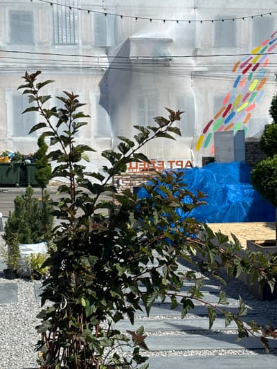 The image captures an outdoor scene in Yelets, Russia, focusing on a building under renovation. A large portion of the building's facade is covered in a white tarp, obscuring its architectural details, though the shapes of windows and balconies are faintly visible. String lights are hung across the front of the building. To the right of the tarp, a colorful, abstract mural of diagonal, elongated shapes in various bright colors is painted on the exposed section of the building.
In the foreground, a dark-leaved plant with multiple branches and serrated leaves dominates the lower half of the frame. The plant is positioned on a pathway paved with large, flat grey stones interspersed with gravel. Behind the plant, to the left, are two green industrial dumpsters, one with trash visible. Further left, a small, conical evergreen tree stands in a white planter.
In the mid-ground, to the right of the plant, a large pile of material is covered with a bright blue tarp. The building behind the blue tarp appears to be constructed of brick, and large orange lettering spelling "APT EMA" is visible on the brick facade. The ground in this area appears to be sand. To the far right, a section of a manicured evergreen hedge is visible. The overall impression is one of an urban setting undergoing construction or modernization, with elements of street art and landscaping present.