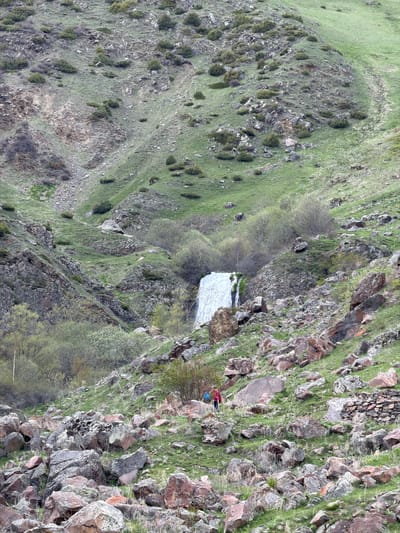 The image depicts a rocky, mountainous landscape in Arsha, Georgia. The terrain consists of steep slopes with patches of grass and sparse vegetation. There's a waterfall visible amidst the rocks and trees. A person is positioned on the right side of the image on a rocky outcrop. They appear to be wearing a red jacket and a blue backpack. The overall environment seems natural, with a rugged, untouched quality.