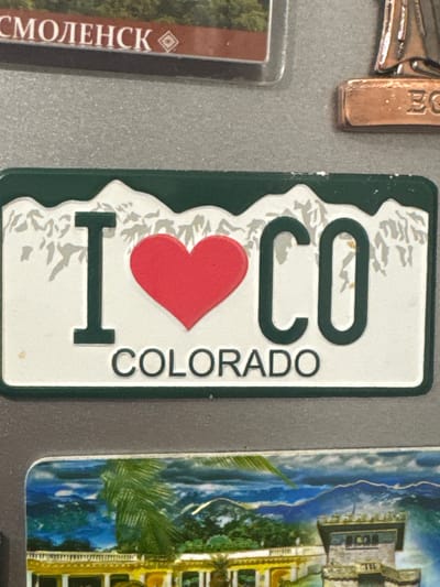 The image is a close-up of a refrigerator magnet. The magnet features a stylized license plate design that says "I COLORADO". The background of the magnet depicts mountains. Above this magnet, there are two other magnets. The one on the left has the text "СМОЛЕНСК" on a red background. The one on the right shows a copper-colored sculpture with the letters "EC" visible. Below the "I COLORADO" magnet is another magnet with a colorful landscape image of mountains and a building that appears to be a castle. The overall surface appears to be a gray, textured wall, possibly a refrigerator door. There are no people visible in the image. The location is not specified in the image.