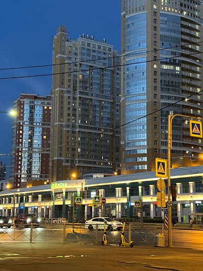 The image depicts a street scene at dusk in Ladozhskoe Ozero, Russia. Tall, modern buildings with illuminated windows dominate the background. In the foreground, a low-rise building with a brightly lit facade features signage with Cyrillic lettering, including "MOMA" in green. Several cars, including a red pickup truck and a white sedan, are visible on the street. Two yellow electric scooters are parked on the sidewalk. Traffic signals and pedestrian crossing signs are present. The sky is a deep blue, indicating twilight. Power lines are strung across the scene. The overall impression is of an urban environment at the beginning of the evening.