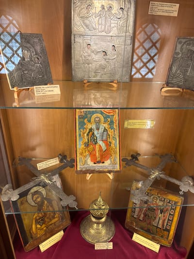 The image shows a display case in the Bachkovo Monastery, Bulgaria, exhibiting religious artifacts. The top shelf features two large, silver relief icons. The left icon, labeled in Russian and English as "ICON, 19th c. s.h. VIRGIN. Jacket: silver.", depicts the Virgin Mary and child. The right icon, also in silver, shows biblical scenes, including a nativity and a head of St. John the Precursor. Below, on the second shelf, is a brightly colored icon of a bearded saint in ornate robes, identified as "Icon of St. John the Baptist, XVIII". Flanking this icon are two large, metal crosses, one with a painted icon of a saint at its center. The cross on the left is labeled "CROSS-SHRINE, 1750. Silver. Gift by Naadji Damaskina". A small, ornate golden censer sits on a red velvet cloth in the foreground, and another icon of Archangel Michael in military attire is visible on the far right. The background is a wooden paneled wall.