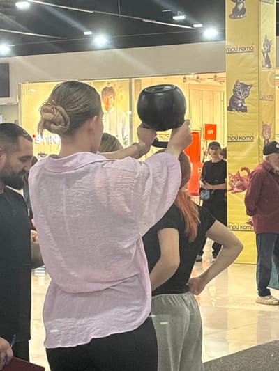 In Tri Protoka, Russia, an indoor scene shows a woman with her hair in a bun, wearing a light purple, textured shirt and black pants, holding a dark-colored kettlebell above her head with both hands. To her left, a man with a beard, wearing a black shirt, is partially visible. To her right, another person with red hair, wearing a black t-shirt and grey pants, stands with their hand on their hip. In the background, a yellow pillar is adorned with images of cats and the text "mou koma." A poster with the number "1599" is also visible, along with other people and displays in the brightly lit indoor environment.