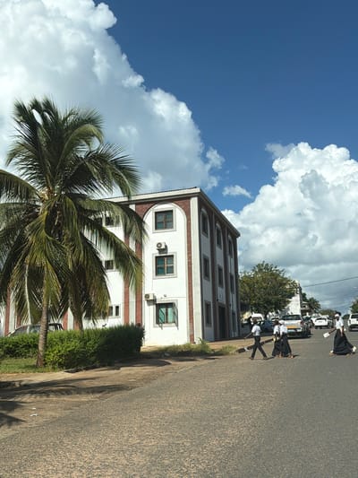 The image presents a partly overcast, daylight scene, possibly in Pemba, Mozambique. A three-story building is positioned along a street; it features a white facade with red brick trim. A tall palm tree dominates the foreground.

On the street, several individuals in what appear to be school uniforms are crossing the road. Some vehicles, including what may be an Isuzu truck, are parked nearby. The paved road exhibits signs of wear. The sky is blue with large, puffy white clouds.