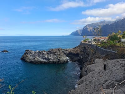 A wide-angle, eye-level shot captures the dramatic coastline of Santiago del Teide, Spain, under a bright blue sky with scattered white clouds. The deep blue sea stretches to the horizon, meeting distant, hazy landmasses. In the foreground, jagged gray rocks form a rugged cliff face, with a prominent, large, dark gray rock formation jutting out into the ocean on the left.
Further along the coast, a paved walkway with a stone wall and railing winds along the clifftop, leading to a cluster of buildings. These structures appear to be part of a resort or residential complex, with white walls, red-tiled roofs, and lush green vegetation, including palm trees, surrounding them. Tall, slender pine trees stand near the buildings, contrasting with the arid landscape. The sheer, dark, rocky cliffs of Los Gigantes rise majestically in the background, their peaks partially obscured by mist. The overall scene is one of natural beauty and coastal development.