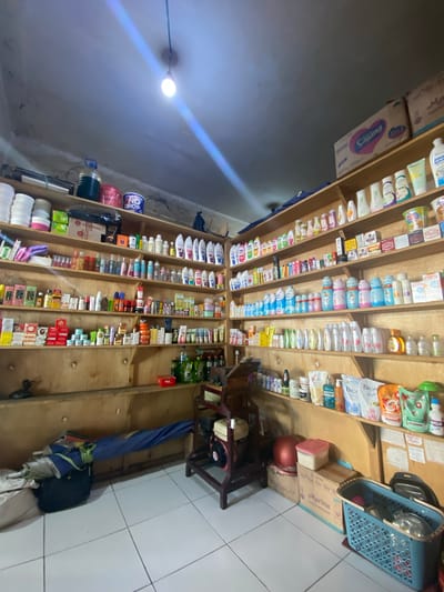 The image shows the interior of a small shop in Ta', Indonesia, stocked with a variety of personal care products and household items. The shop features wooden shelves filled with products such as lotions, soaps, shampoos, and cleaning supplies. There are also some boxed goods and a few larger containers. In the foreground, a blue basket containing various items is visible, along with a few storage boxes and a red stool. The lighting in the shop appears to be from an overhead bulb. The floor is tiled, and the walls are made of concrete. The overall impression is of a well-stocked local convenience store or pharmacy.