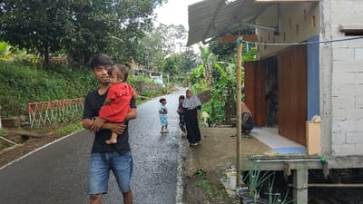 This image captures a lively, casual street scene in a rural or semi-rural residential area, likely in Kecamatan Panjalu, Indonesia.

In the left foreground, a young man with short dark hair, wearing a black t-shirt and denim shorts, holds a baby dressed in a bright red outfit. He smiles gently as he looks down at the baby, who appears calm and gazes towards the right. They stand on a narrow, wet paved road that recedes into the background.

To the left of the road, a steep, verdant hillside is covered in dense green foliage and trees, with a red and white painted metal barrier or railing running along its base. Further down the road, closer to the center of the image, two young children are visible. One child wears a blue and white patterned outfit, and the other a dark shirt. They stand near a woman dressed in a black abaya and a grey hijab. This group appears to be interacting or observing the scene.

On the right side of the image, a simple house or building with unfinished cinder block walls and wooden sliding doors is partially visible. A makeshift lean-to roof extends over the entrance. A person, possibly a woman, is seen crouching near the entrance of this building, seemingly engaged in a task. Several potted plants, including some with tall green leaves resembling spring onions, are arranged on a ledge beneath the building. Lush tropical vegetation, including large-leafed plants like banana trees, grows around the structures.

The road's wet surface suggests it may have recently rained. The lighting is soft and natural, indicating daytime, possibly under an overcast sky or during the early morning/late afternoon. The scene conveys a sense of community life amidst abundant greenery. There is no visible text in the image.