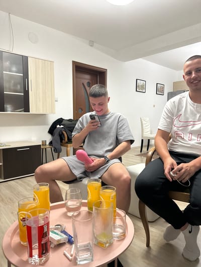 The image depicts two men in an indoor setting. One man is sitting on a chair wearing black pants and a white shirt with a Nike logo. He is holding something in his hands. The other man is in the foreground wearing a grey t-shirt and shorts, looking at a mobile phone and holding a pink object. A round table with glasses and cigarettes is also in the foreground. The location appears to be inside a home or apartment. The city of Graf Ignatievo, Bulgaria is not identifiable from the image.