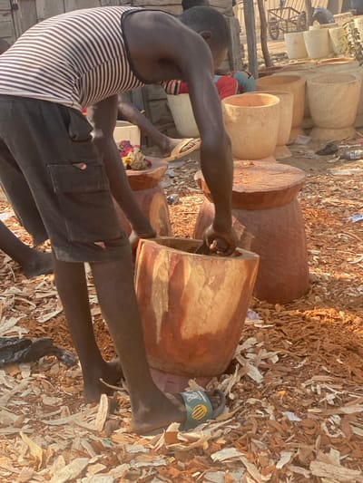 In Zongon Daji, Nigeria, an outdoor scene depicts a dark-skinned individual engaged in woodworking. The person, positioned in the foreground and facing partially away from the camera, wears a white and dark-striped sleeveless tank top and dark cargo shorts. A dark-green slide sandal with a white logo is on their right foot, while their left foot is bare. They are bent over a large, reddish-brown wooden mortar, actively hollowing out its interior with a tool held in their right hand. The ground around them is thickly covered with light-colored wood shavings and chips.
To the right of the primary subject, another reddish-brown wooden object, possibly a stool or a different type of mortar base, stands on the ground. In the midground, several light-colored, finished or semi-finished wooden mortars are visible, arranged in rows. Further back, another person in red and blue clothing is partially visible, seated or crouched. The background includes a light-colored building structure, a cart, and sparse vegetation under a bright, possibly sunny, sky, indicating an outdoor workshop or market setting.