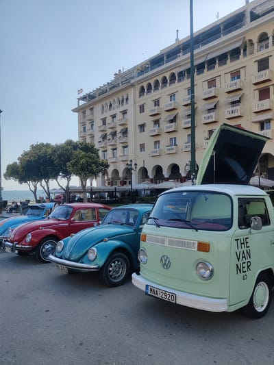 In Thessaloniki Municipal Unit, Greece, a row of vintage Volkswagen cars is parked along a street next to the sea. The cars, including several iconic Volkswagen Beetles in blue and red, and a light green Volkswagen Type 2 van with its top open, are aligned facing away from the viewer. The van has the words "THE VANNER S" printed vertically on its side. Behind the cars, a large, light-colored building with arched balconies and awnings dominates the background. Trees and outdoor seating areas for a cafe are visible between the cars and the building. The sky is clear and blue, and the overall atmosphere suggests a pleasant, sunny day.